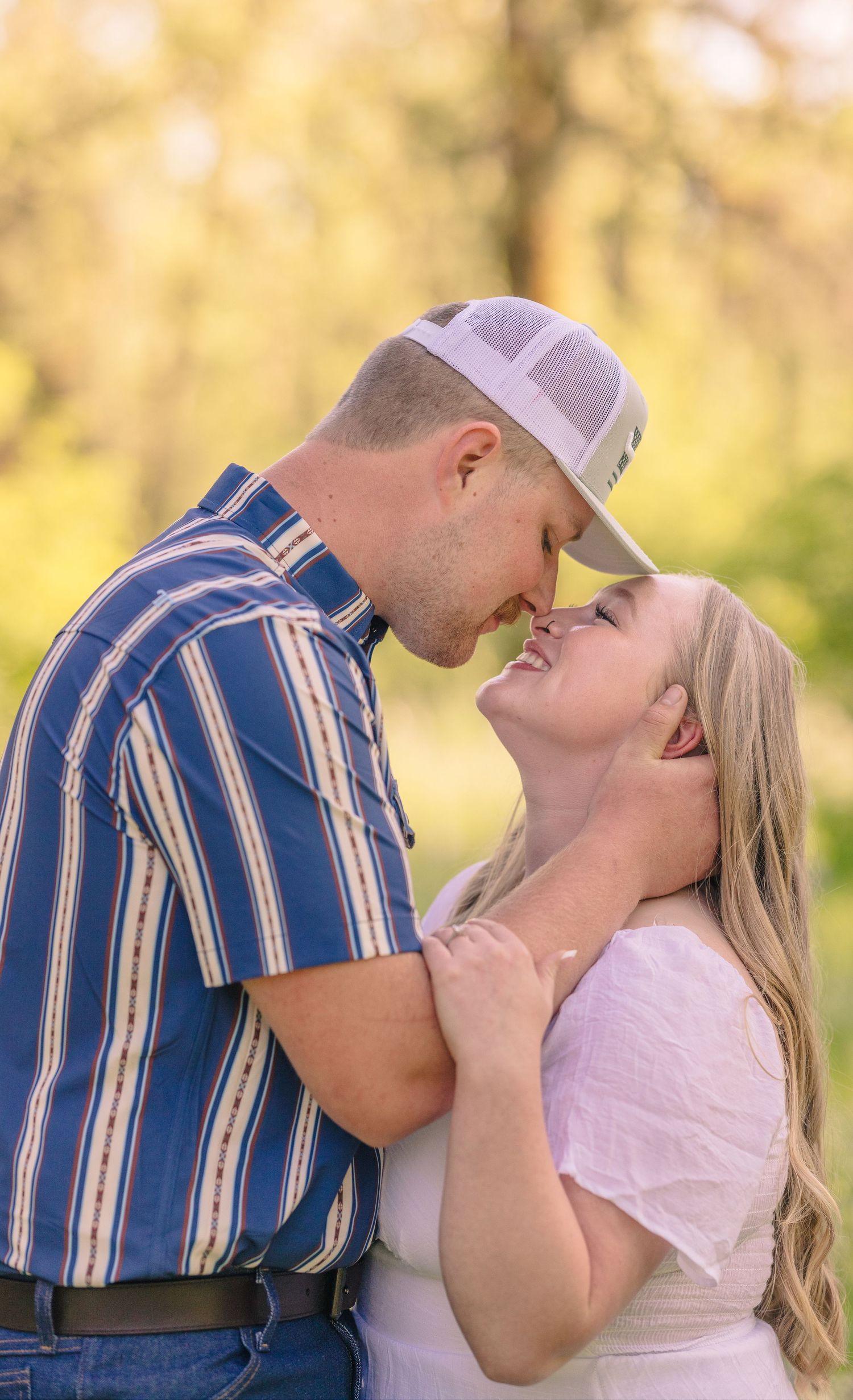 Couple sharing a romantic moment outdoors, man in striped shirt and white cap leaning in to kiss smiling blonde woman.