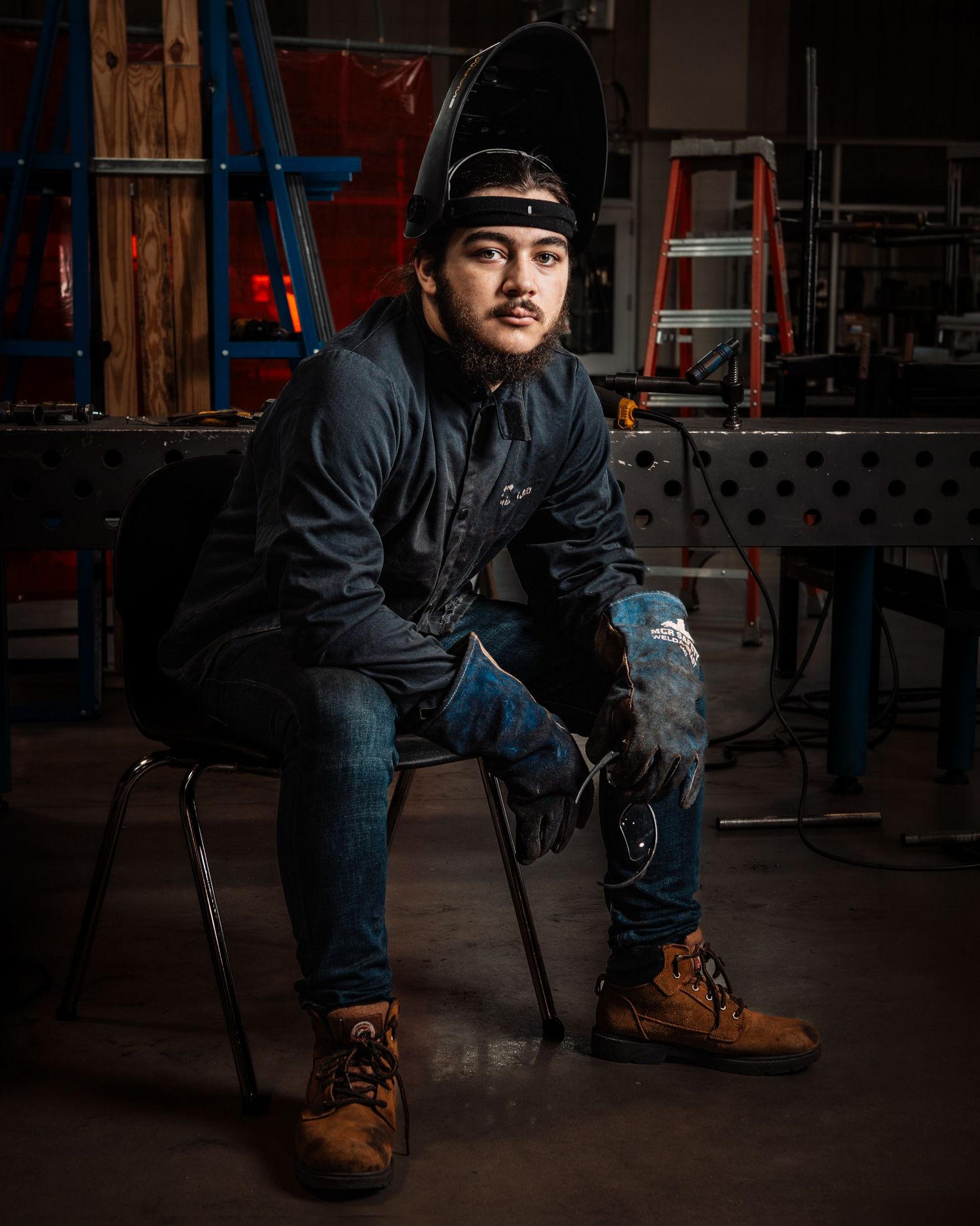 A young welder sits in a workshop chair wearing a welding helmet, dark jacket, jeans, and brown boots.