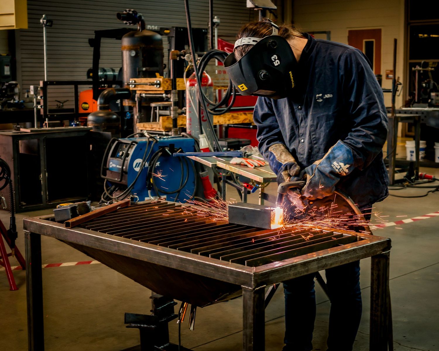 A welder in protective gear creates sparks while working on metal grating in an industrial workshop.