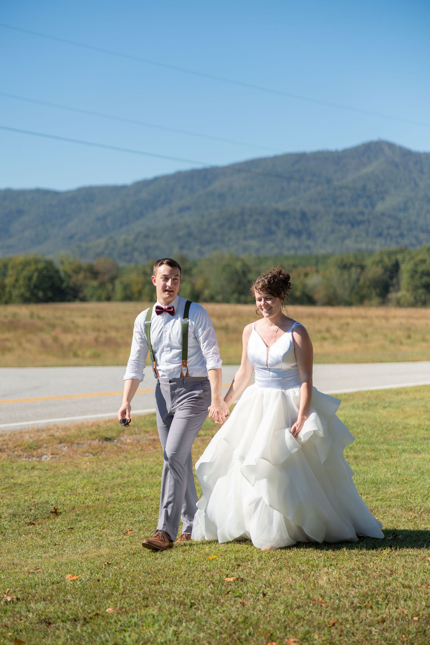 A bride and groom walk hand-in-hand across a green field with mountain scenery under a clear blue sky.