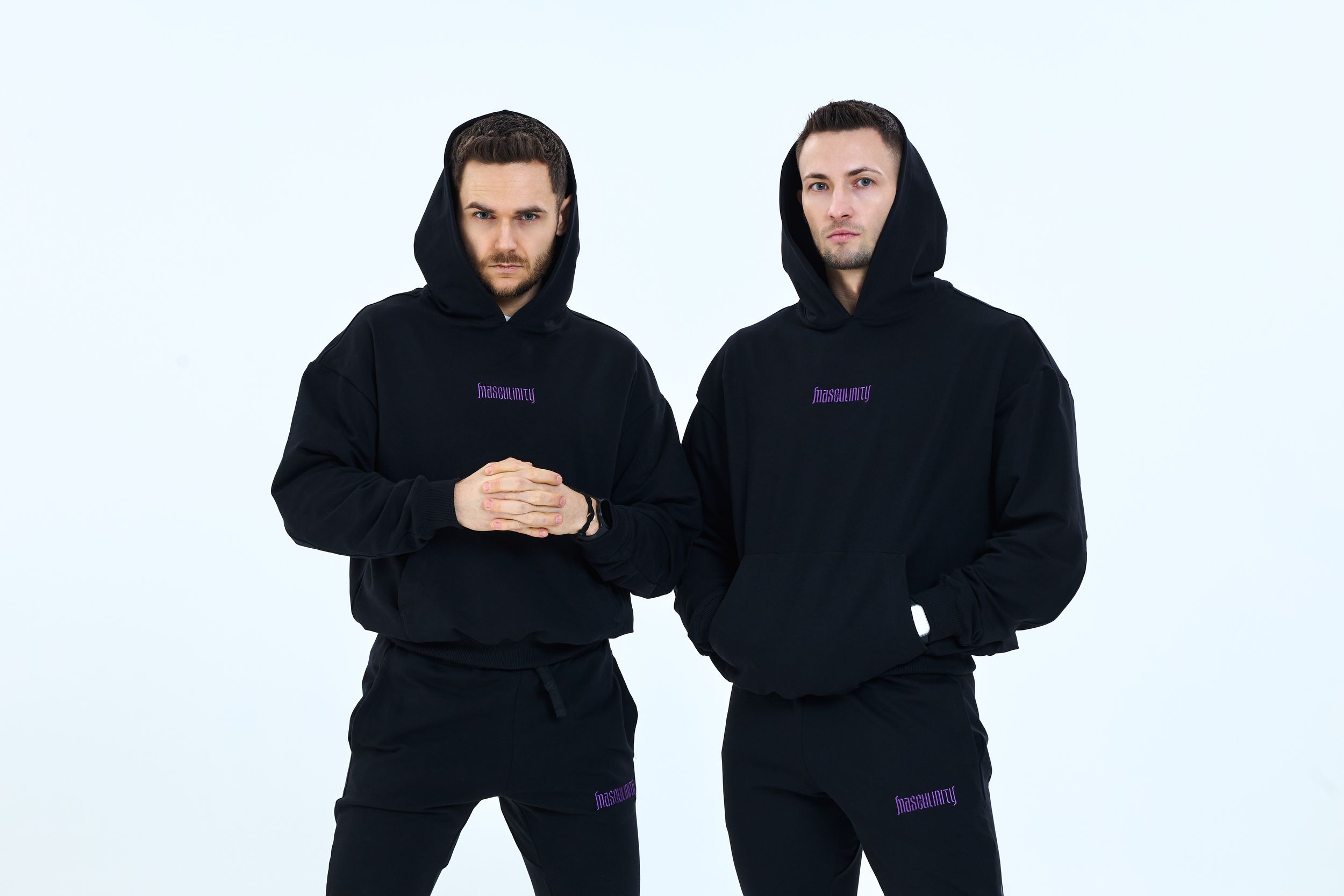 Two men wearing matching black hoodies and sweatpants with purple logo print, posing against a white background.