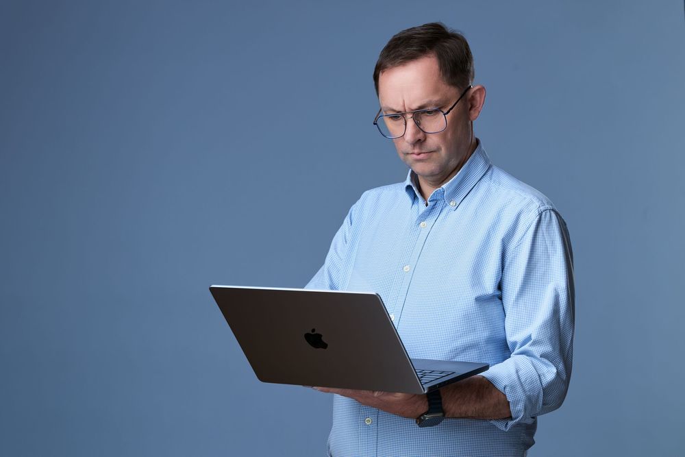 Middle-aged man in blue shirt and glasses holding an Apple MacBook laptop against a blue background.