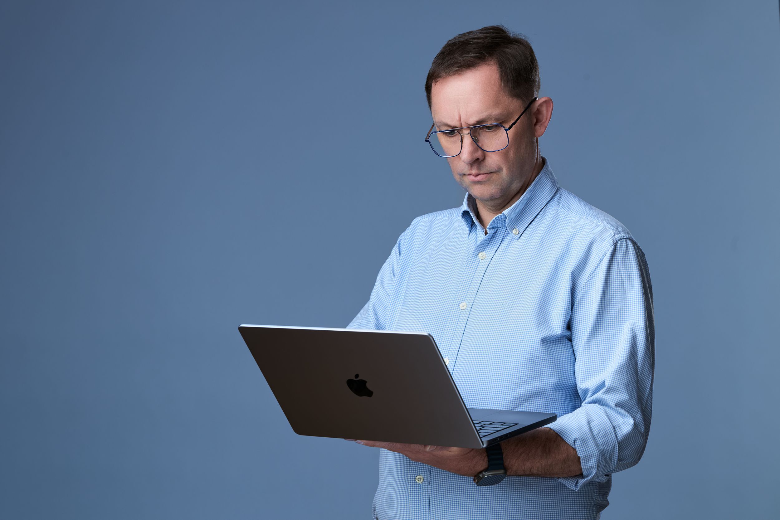 Middle-aged man in blue shirt and glasses holding an Apple MacBook laptop against a blue background.