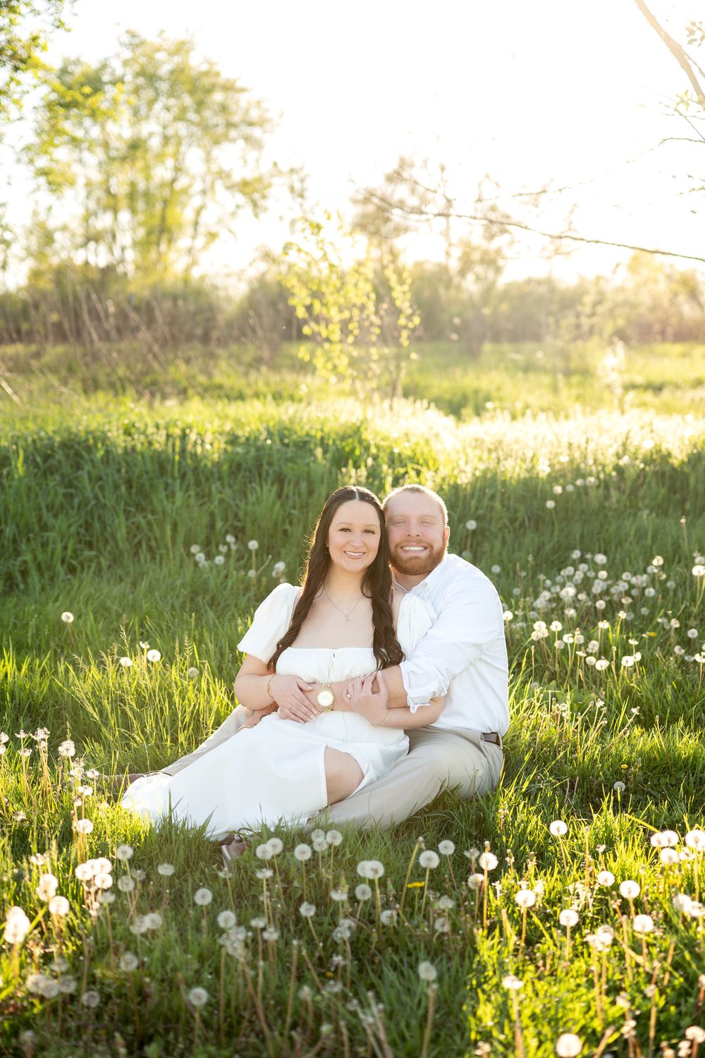 Smiling couple sitting together in a sunlit meadow filled with white dandelions.