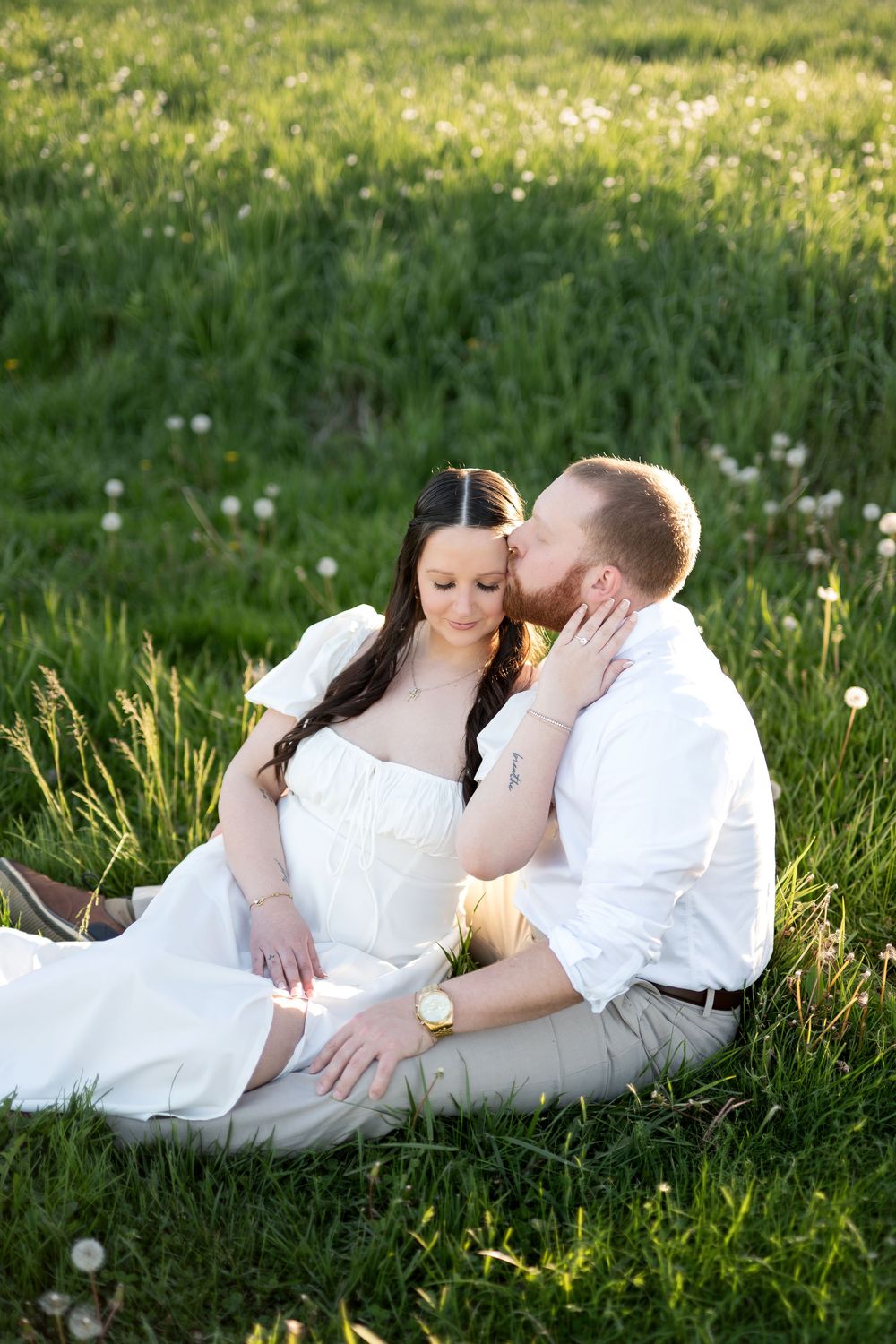 Man kissing woman on cheek while sitting together in a sunlit green dandelion meadow.