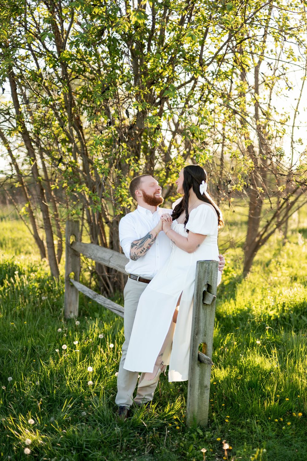 Couple embracing by rustic fence in lush green field at golden hour.