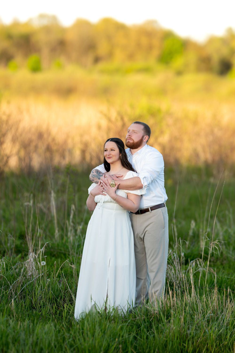 Couple embracing in golden field at sunset, man in white shirt hugging woman in flowing white dress.