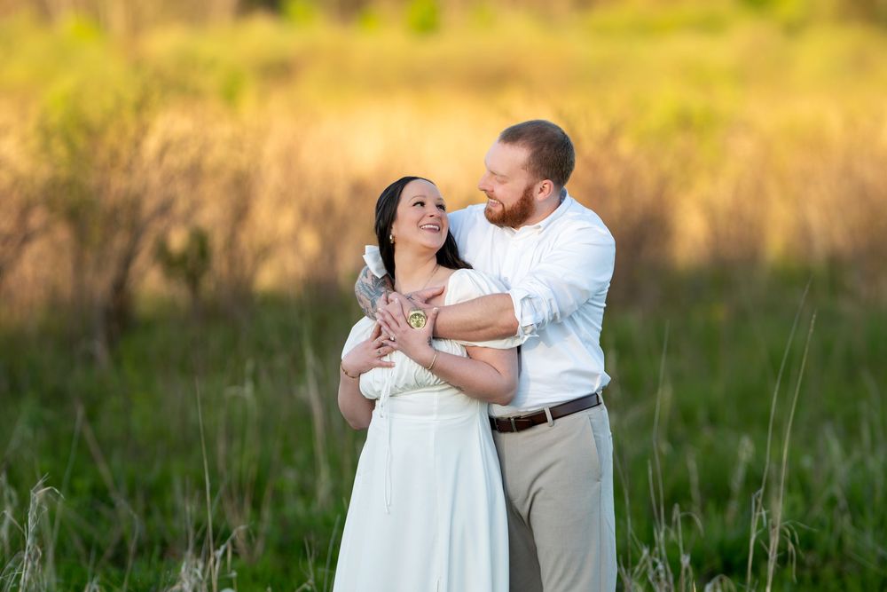 Smiling couple sharing a tender moment in a sunlit meadow, woman in white dress held by bearded man.