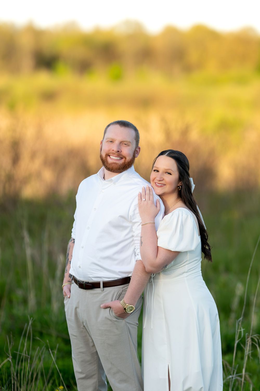 Happy couple posing in a sunlit field, woman in white dress leaning against smiling bearded man.