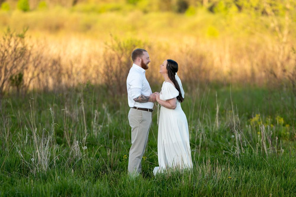 Couple facing each other lovingly in a green field at golden hour, woman in white dress.