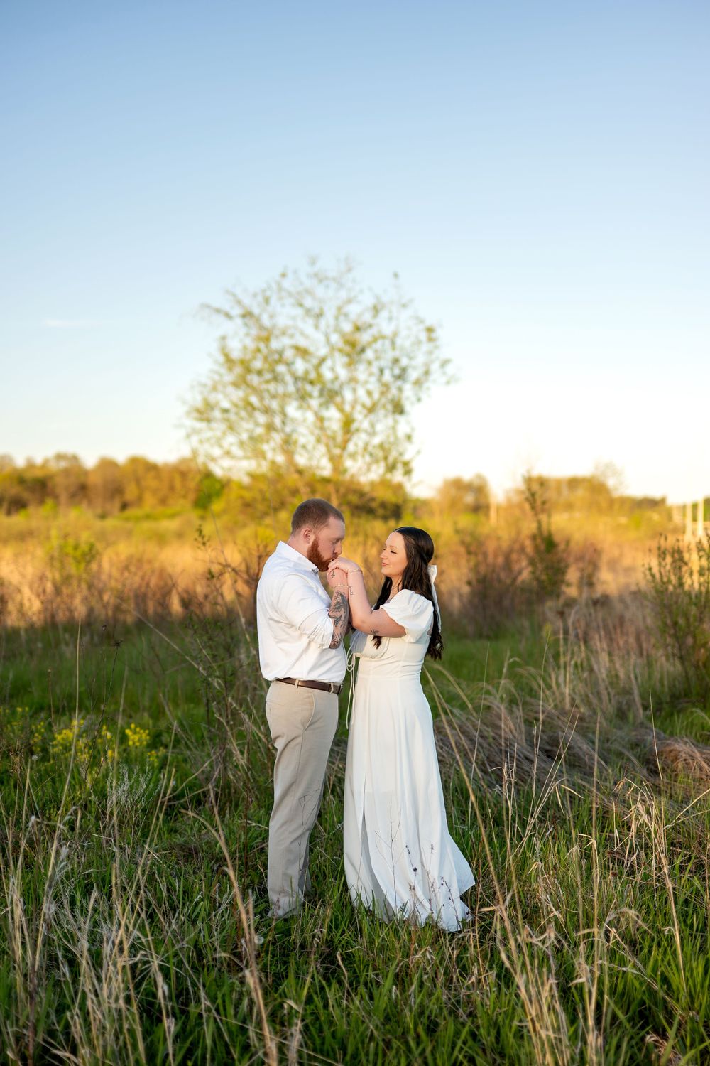 Bearded man kissing woman's hand as she smiles in a white dress during outdoor engagement photos at sunset.