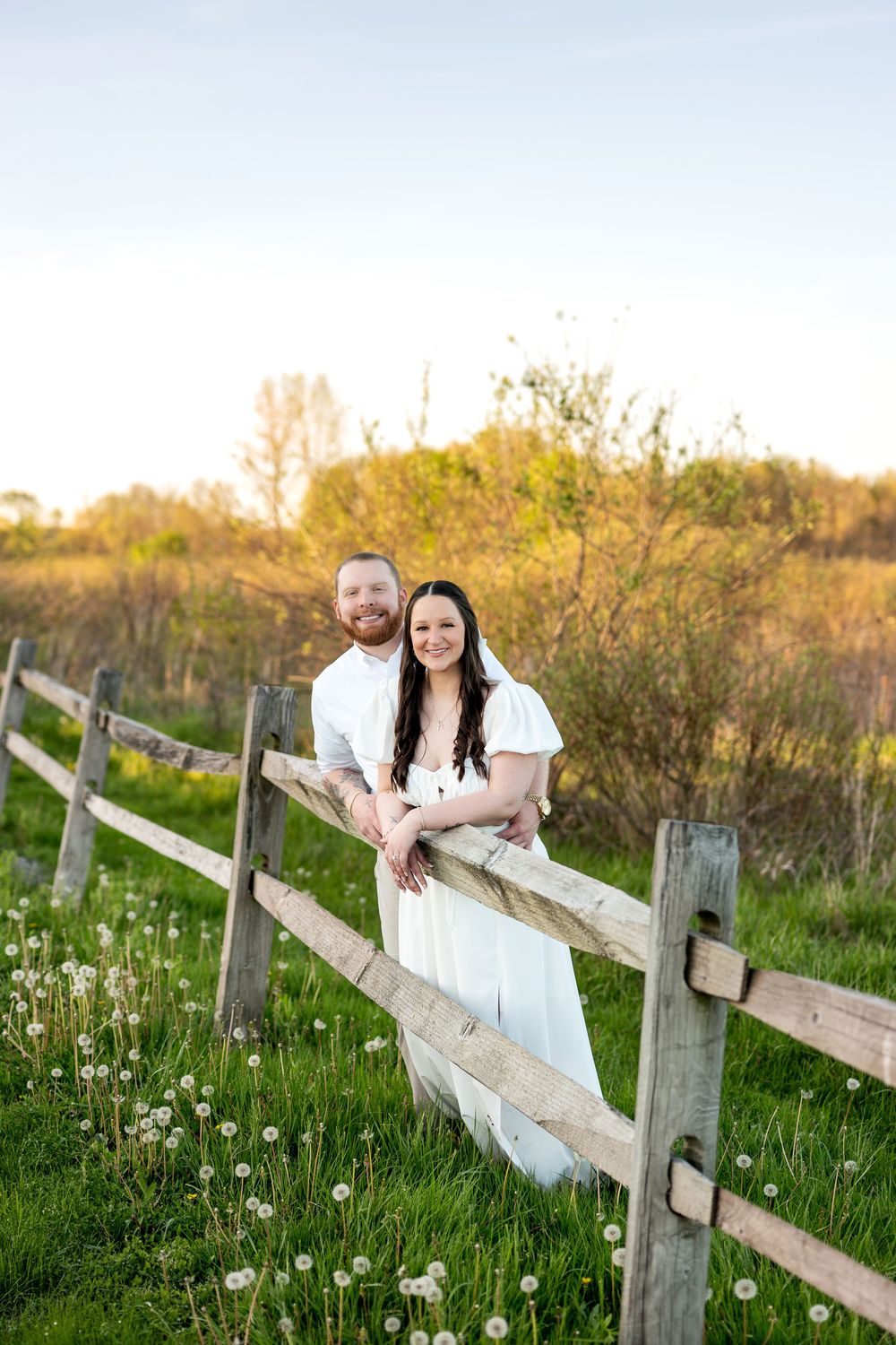 Happy couple posing together along a split-rail fence in a green meadow at sunset.