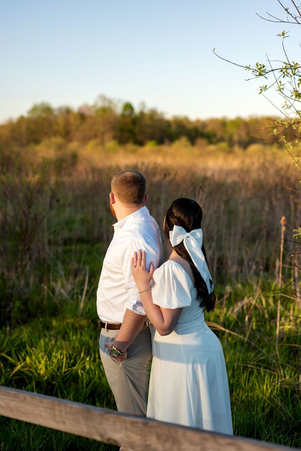 Couple standing by a wooden fence overlooking a sunlit meadow, woman wearing large white bow in her hair.