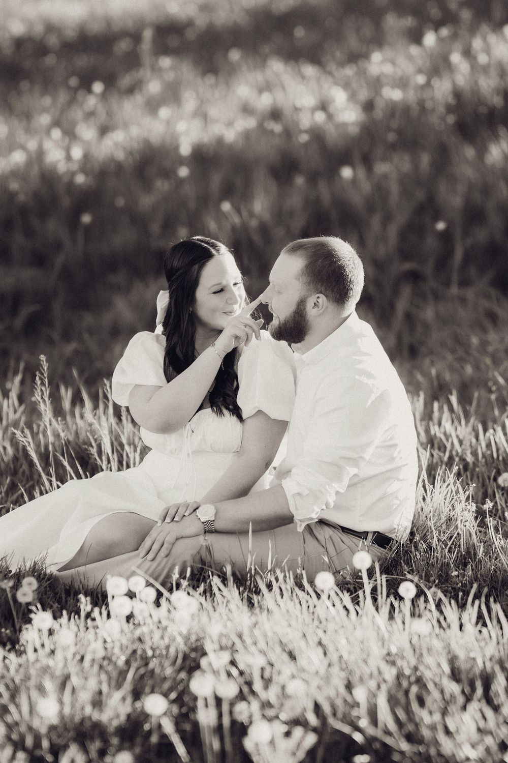 Black and white photo of a couple sitting in a dandelion field, woman touching mans nose affectionately.