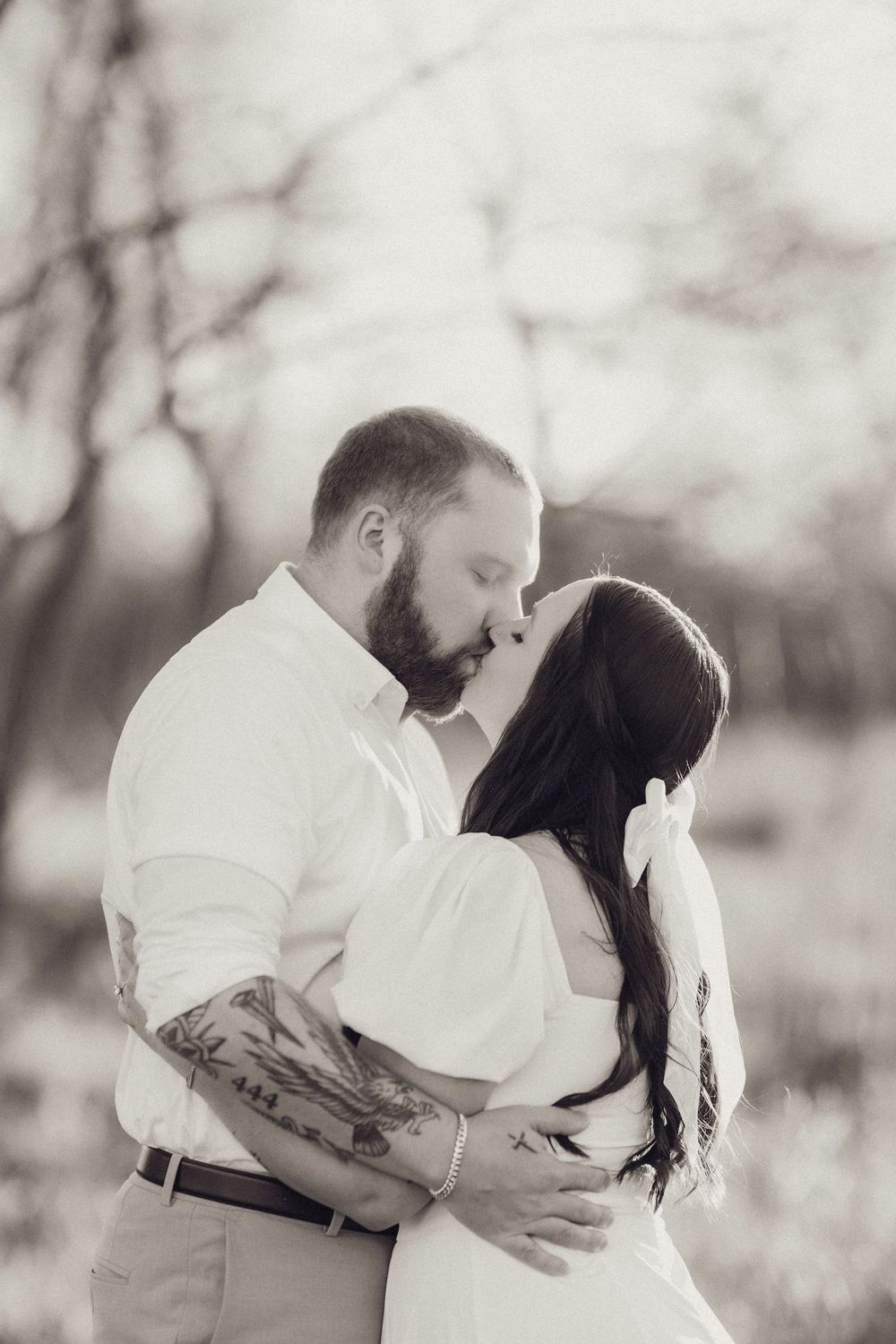 Black and white photo of a tattooed couple sharing a romantic kiss outdoors among bare trees.