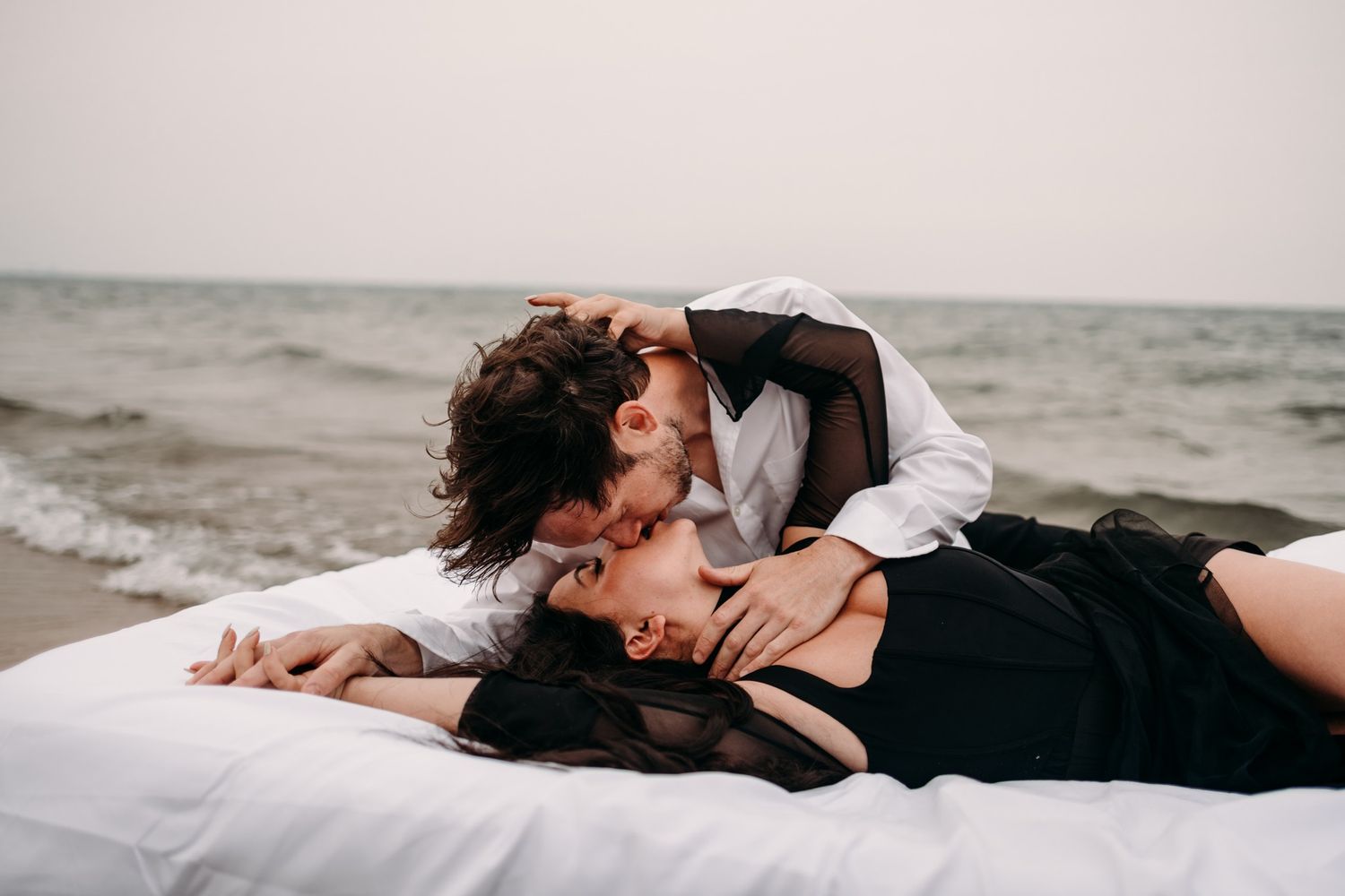 Romantic couple kissing on white bedding at the beach, woman in black dress, man in white shirt, ocean backdrop.