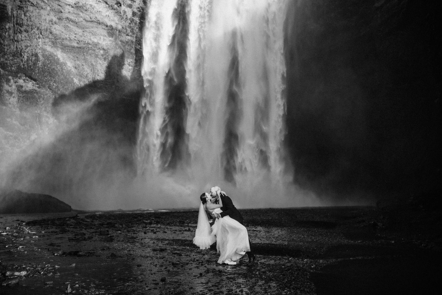 Black and white photo of a bride kneeling before a massive waterfall in Iceland, dress flowing dramatically.