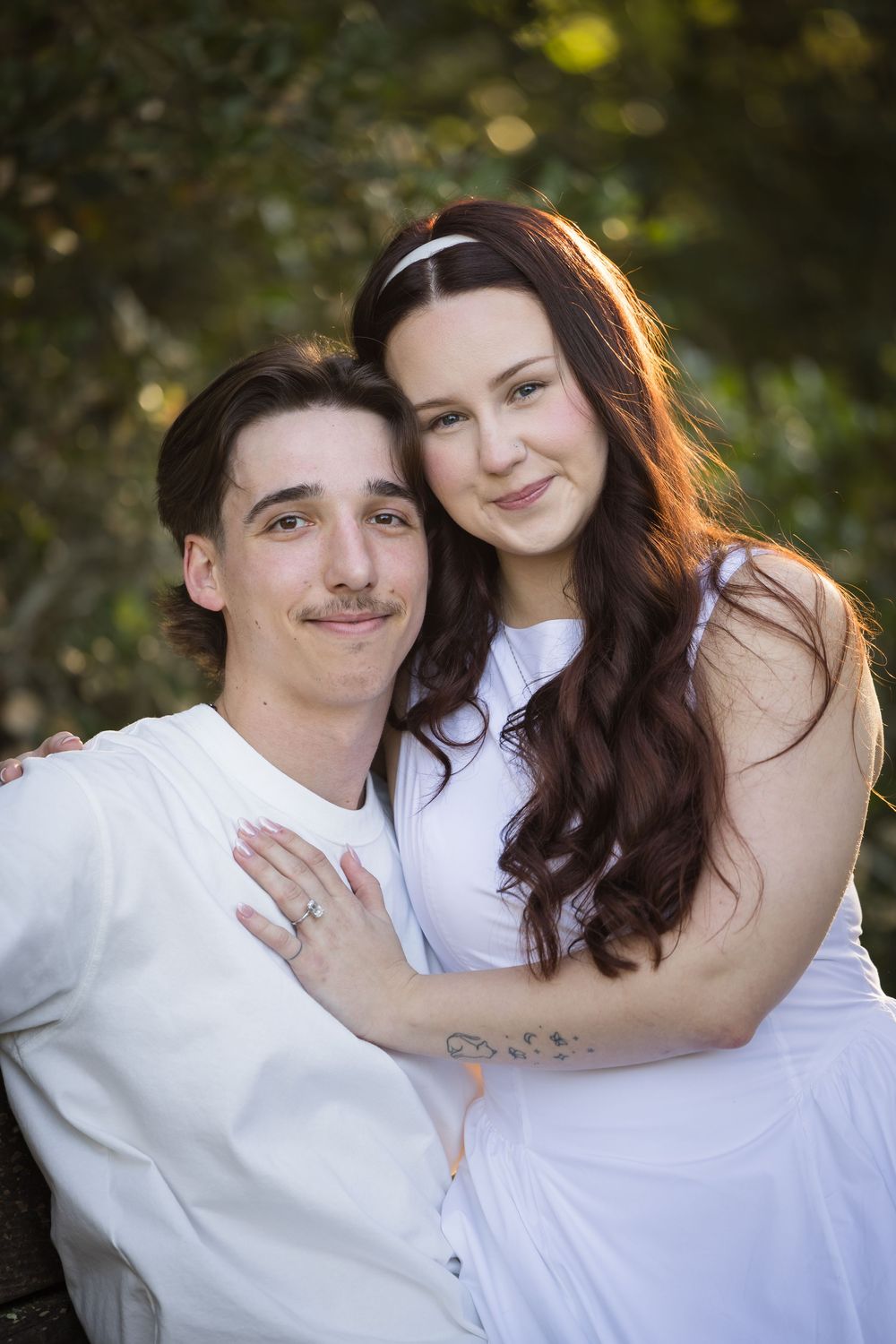 Romantic couple in white outfits sitting together outdoors, woman with long wavy hair resting on mans shoulder.