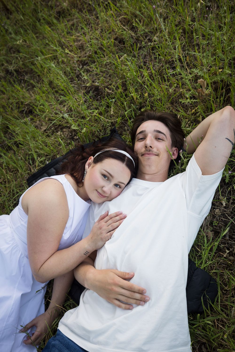 A young couple lies on green grass, the woman resting her head on the mans chest affectionately.