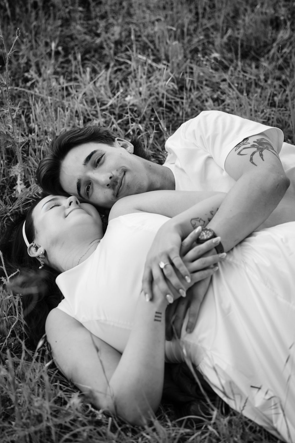 Black and white photo of a couple lying in grass, embracing tenderly with closed eyes.