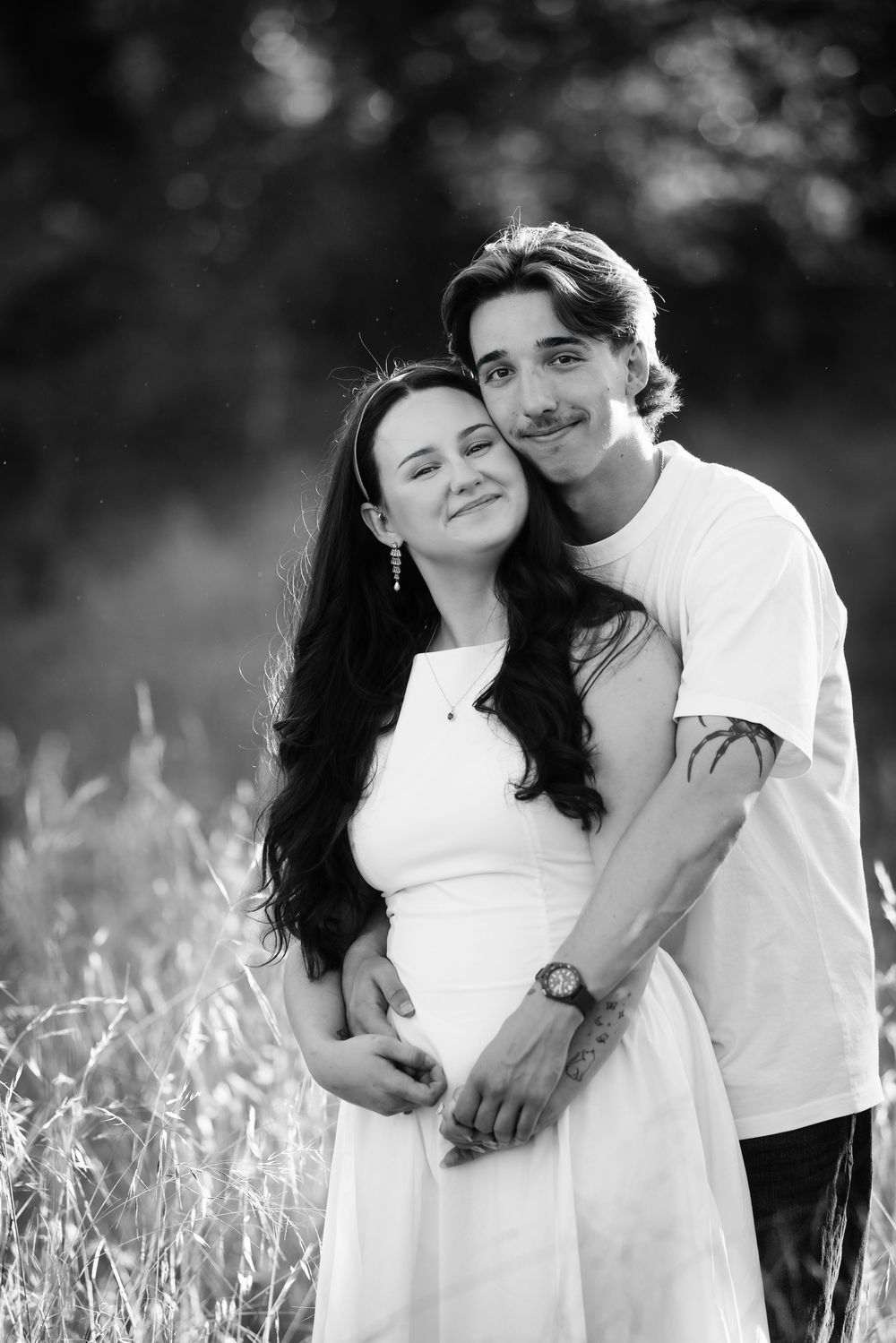 Black and white photo of a young couple embracing in a field, woman in white dress smiling peacefully.