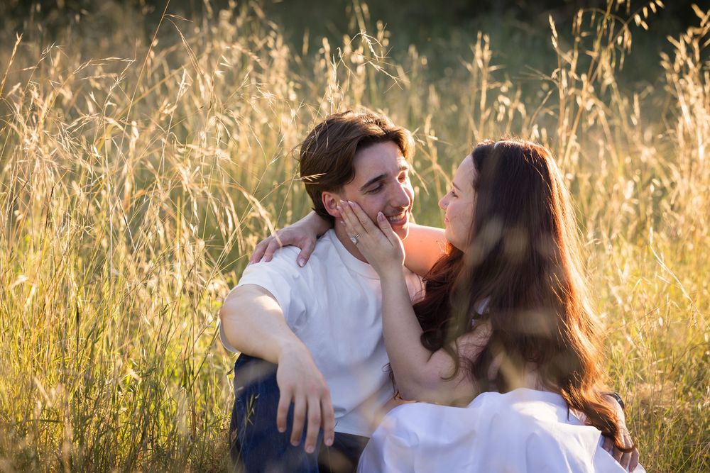 Romantic couple sitting in a sunlit golden field, gazing at each other affectionately in warm light.