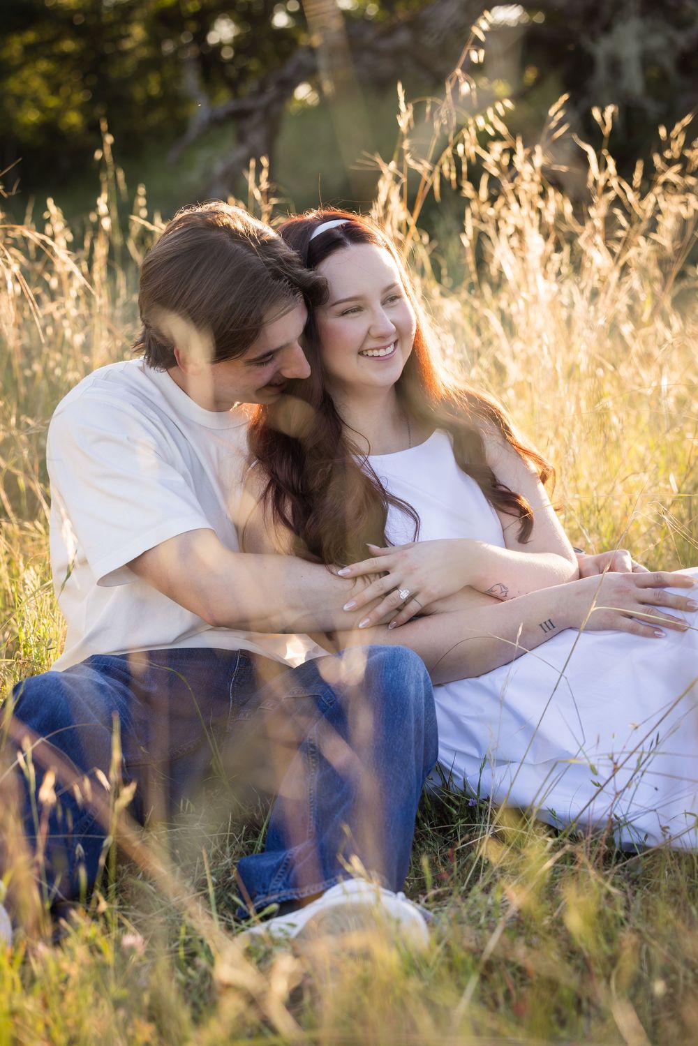 Couple sitting in golden grass field, embracing and smiling, wearing white tops and jeans in warm sunlight.