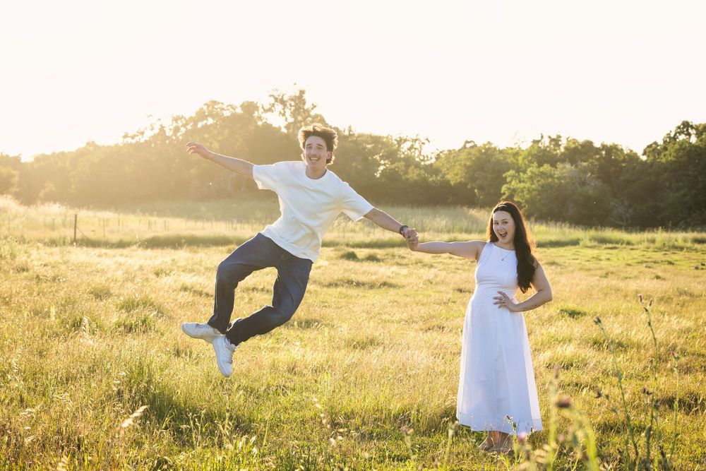 Joyful couple in a sunny field, man jumping while holding hands with woman in white dress.