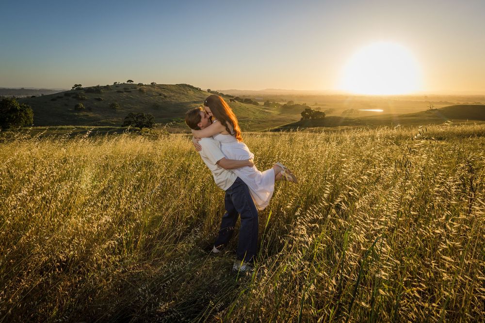 Couple embracing in golden field at sunset, man lifting woman in romantic outdoor engagement photo.