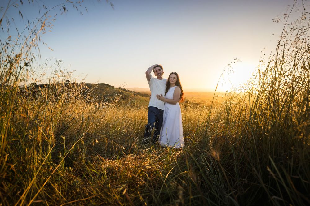 Couple embracing in golden field at sunset, man raising arm overhead, woman in white dress, warm sunlight glowing behind them.