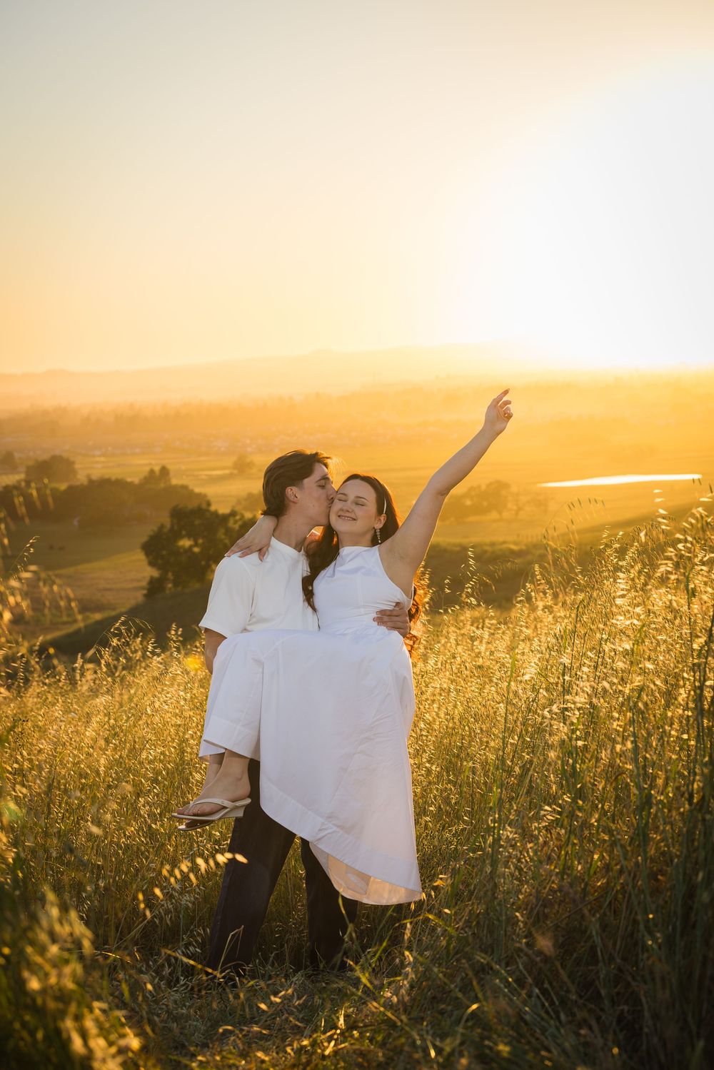 Couple in white embracing at golden sunset in a grassy field, woman reaching up joyfully with scenic valley backdrop.
