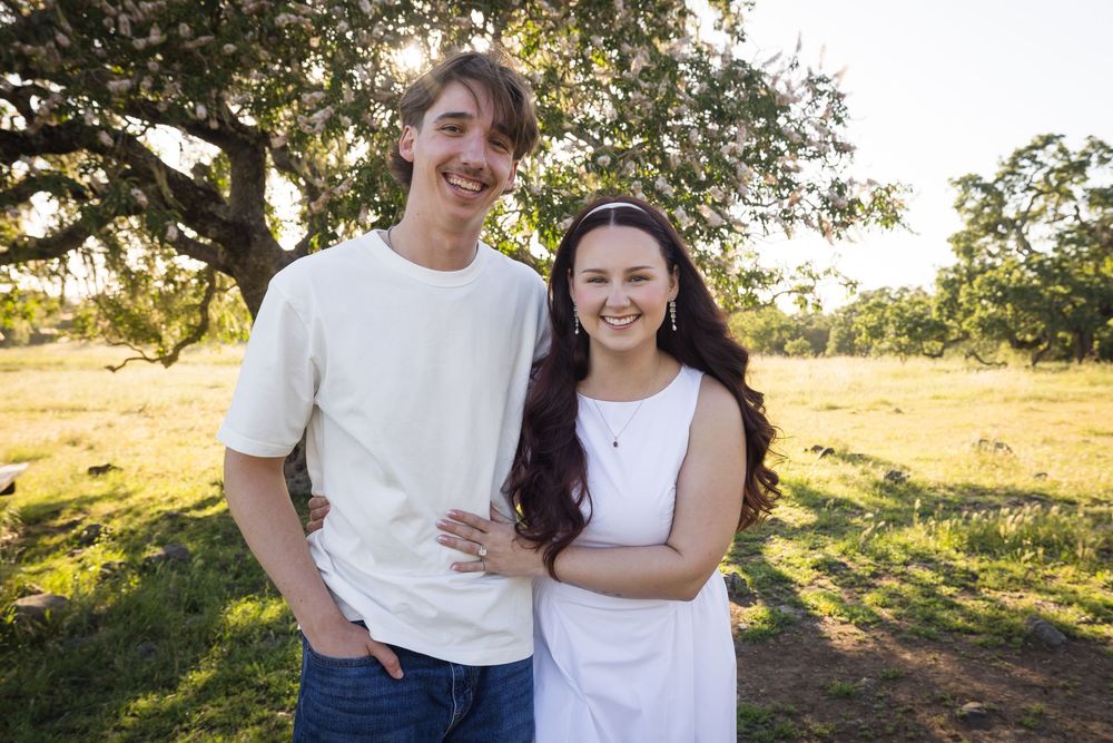 A young couple smiles outdoors under a tree in a sunny green field, wearing white outfits.