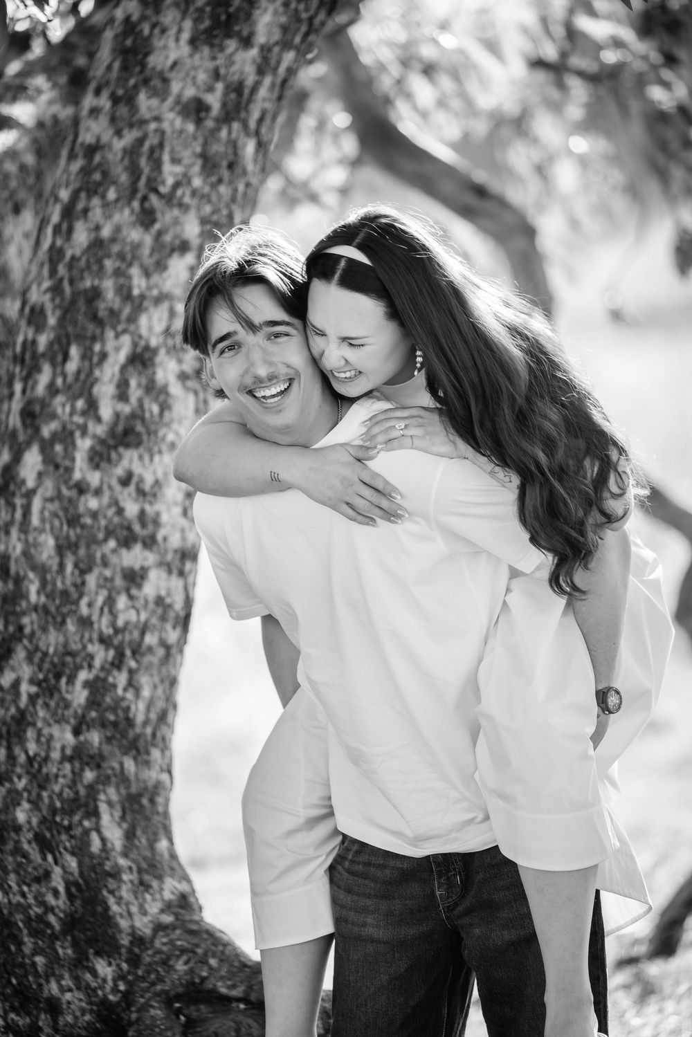 Black and white photo of a happy couple, man giving woman a piggyback ride while smiling outdoors near a tree.