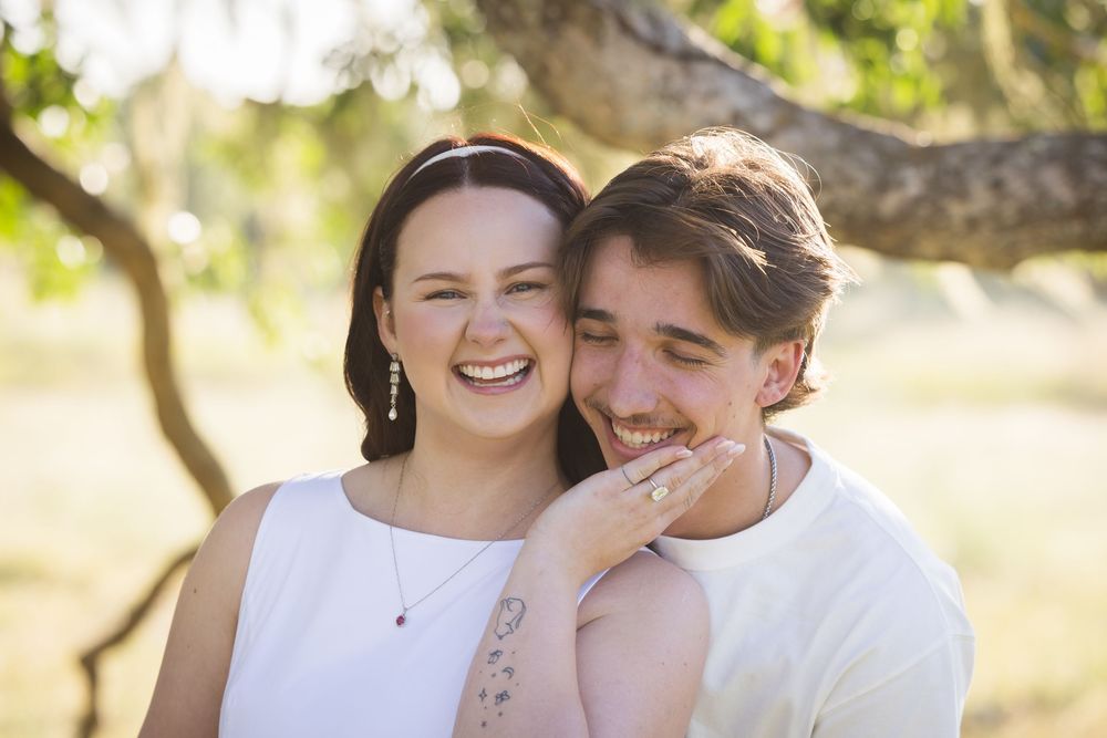 Happy young couple laughing together outdoors in a sunny park, woman in white dress with hand on mans shoulder.