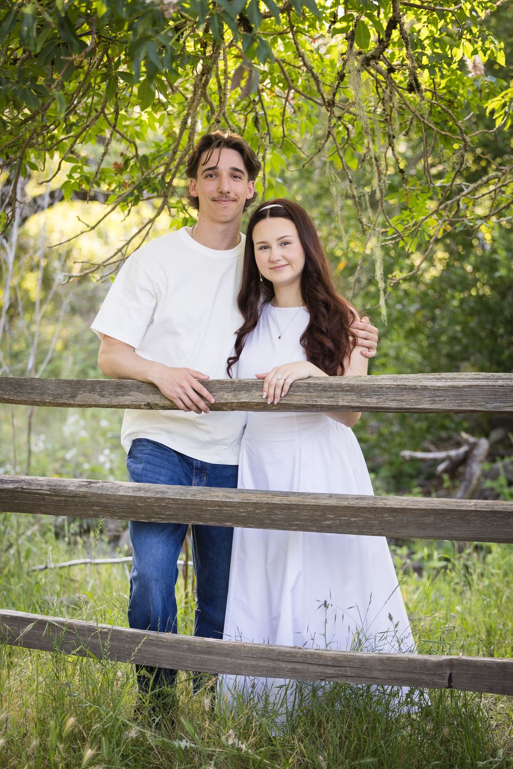 Young couple in white outfits smiling by a rustic wooden fence surrounded by lush green trees outdoors.