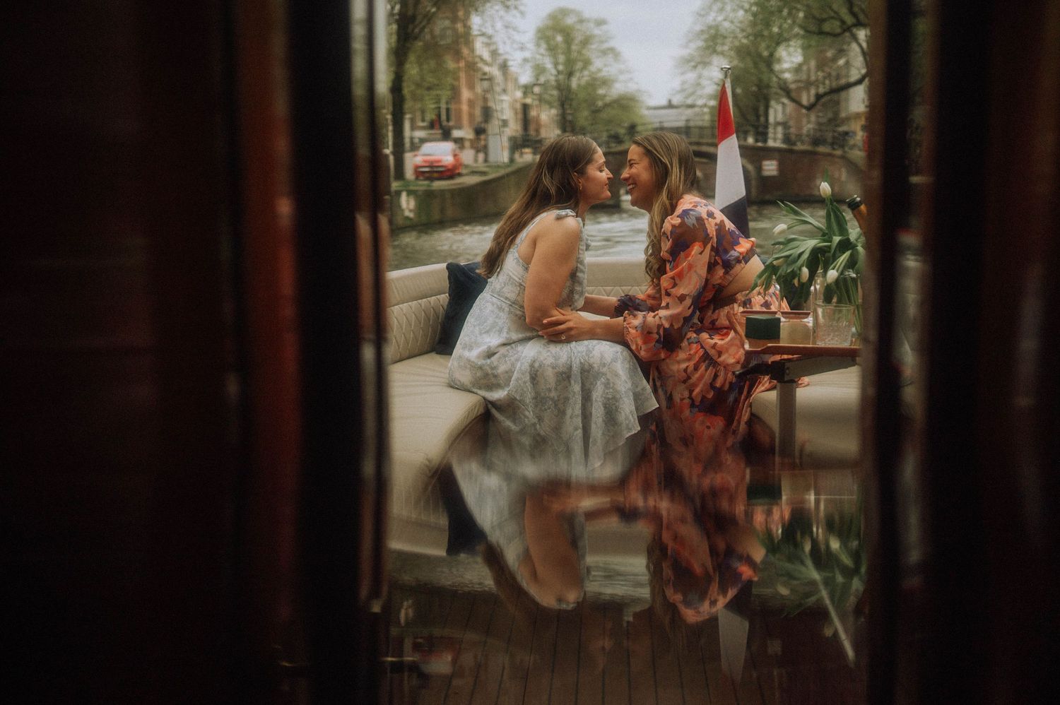 Two women share a romantic moment sitting together on a canal boat in Amsterdam during secret proposal photoshoot, reflected in a window.