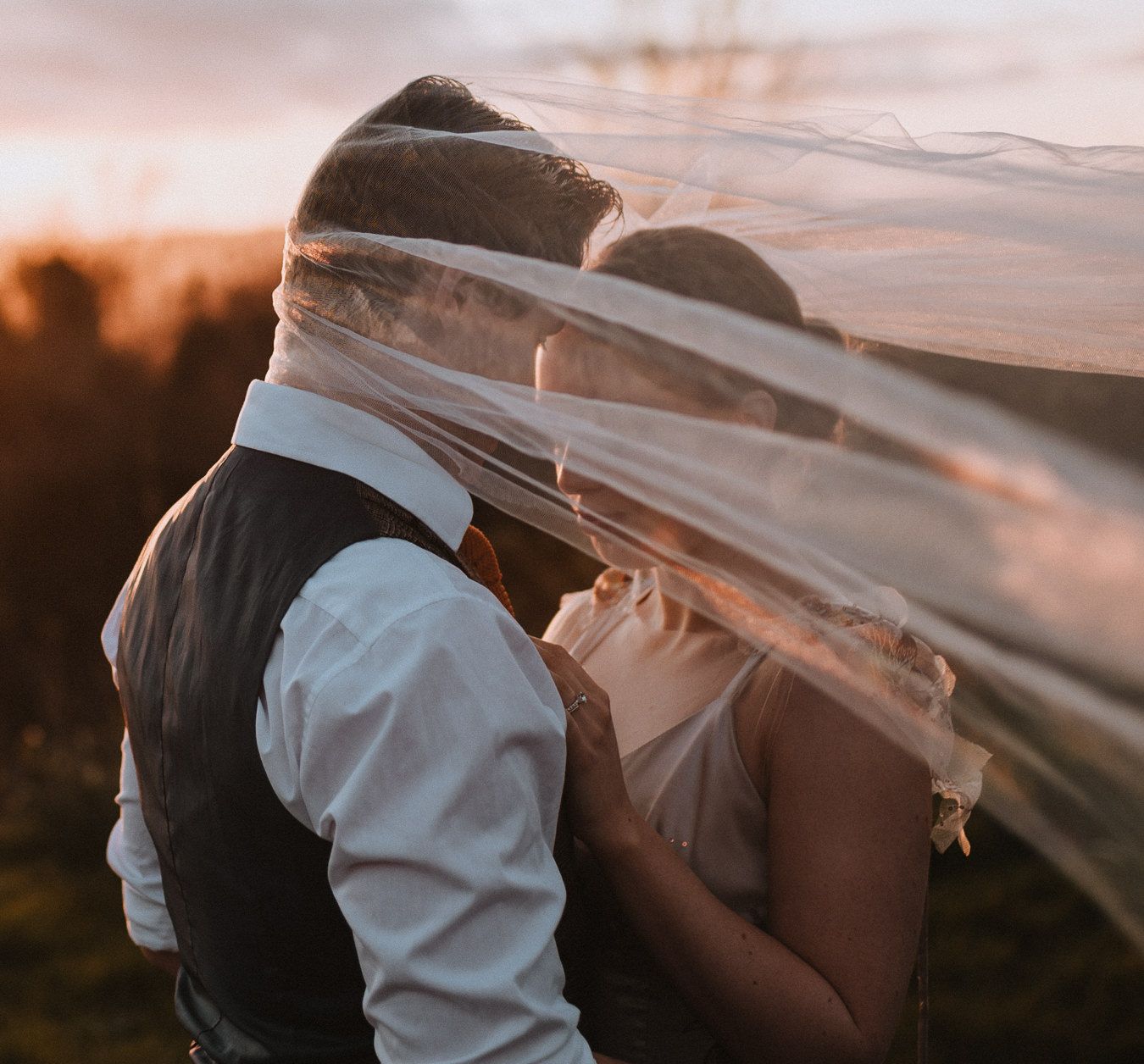 A bride and groom share a romantic kiss under a flowing wedding veil at golden sunset.