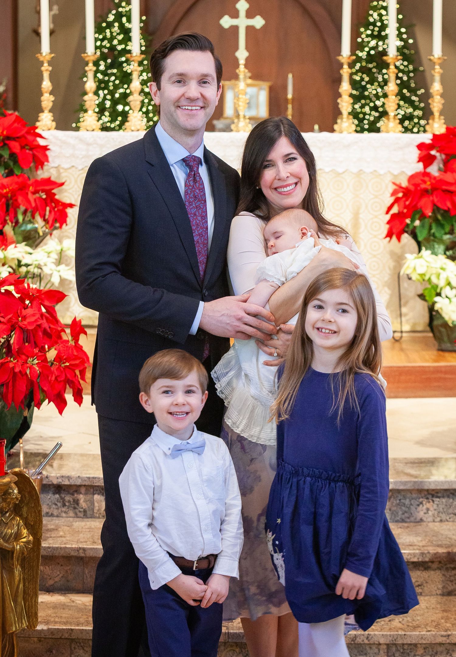 Happy family of five poses at church altar decorated with red poinsettias for Christmas baptism.
