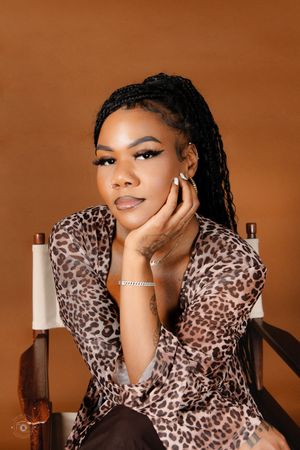Woman in leopard print outfit with box braids poses gracefully in a wooden chair against a warm brown background.