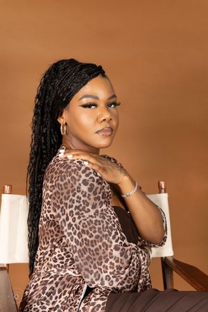 A woman with long black braids wearing a leopard print top sits in a director's chair against a brown background.