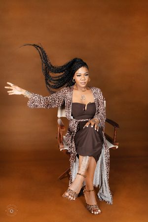 Woman in leopard print robe and brown dress poses dramatically in chair with braids flowing, studio portrait.