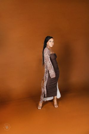 Pregnant woman in brown bodycon dress and leopard print duster coat poses against warm brown studio background.