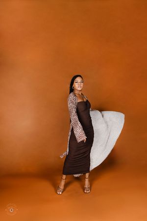 Woman in brown midi dress and leopard cardigan poses gracefully against warm orange background holding white fluffy pillow.