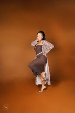 Woman posing in brown midi dress and leopard print cardigan with silver heels against warm brown studio background.