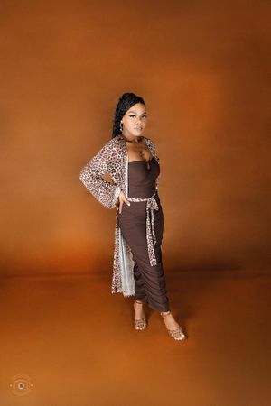 Woman posing in brown ruched dress with leopard print cardigan and clear heels against orange background.