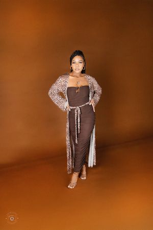 Woman in brown midi dress and leopard print cardigan posing confidently against warm brown studio backdrop.