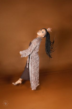 Woman in leopard print kimono with long braids poses dynamically against a warm brown studio background.