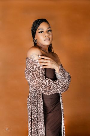 Woman in leopard print cardigan and brown dress poses confidently against warm orange studio background.