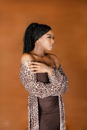 Woman in leopard print cardigan and brown dress with braids and tattoos posing against orange background.