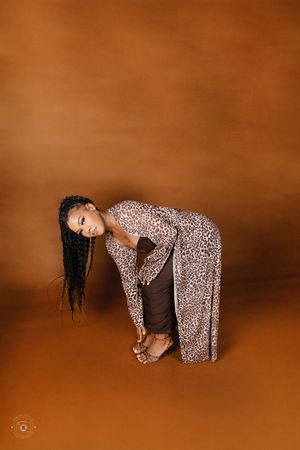 Woman in leopard print cardigan bends gracefully toward floor against warm amber background.